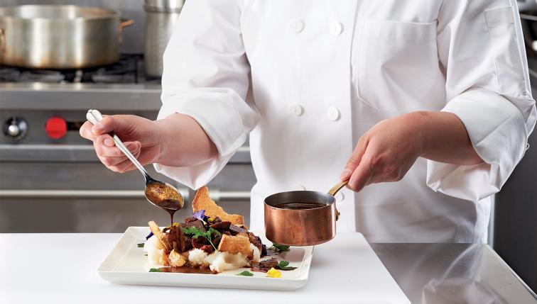 A chef uses a spoon to top a beef dish with gravy.